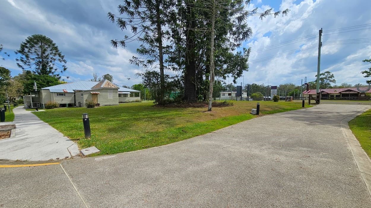 A grassy area with a large tree in the centre. On the left, there's a light-coloured house with a metal roof and a concrete path leading towards it. On the right, a paved path curves through the grass, bordered by small black bollards. In the background, there are more houses and utility poles with wires. The sky is partly cloudy.