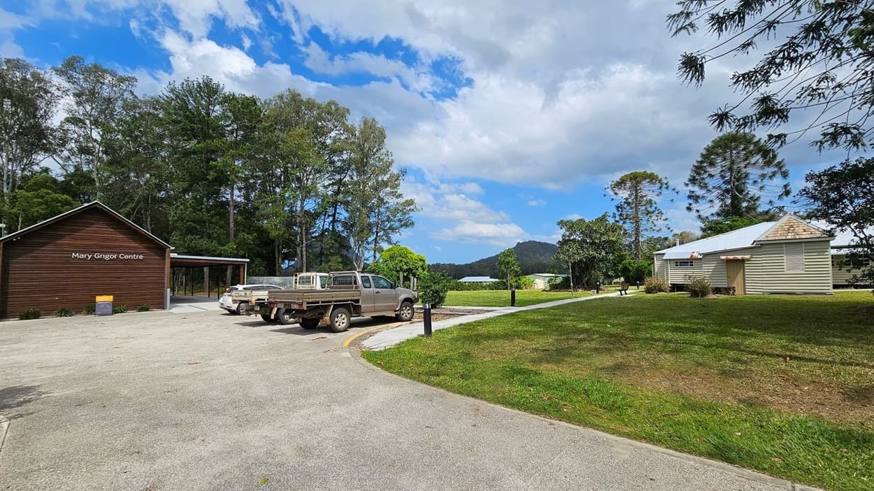 A rural scene with a wooden building on the left labelled "Mary Grigor Centre" surrounded by tall trees. Two utility vehicles are parked nearby. On the right, a small, light-coloured house with a metal roof sits on a grassy area. A path runs between the buildings, and there are trees and a mountain in the background. The ground is a mix of grass and paved surfaces.