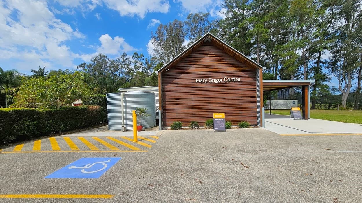 A building with a wooden facade labelled "Mary Grigor Centre" is in the centre. To the left, there's a large metal water tank and a hedge with trees behind it. In the foreground, a parking area features a blue disabled parking sign and yellow-striped markings. On the right, a covered area extends from the building, with trees and grass in the background. A small sign is placed near the entrance.