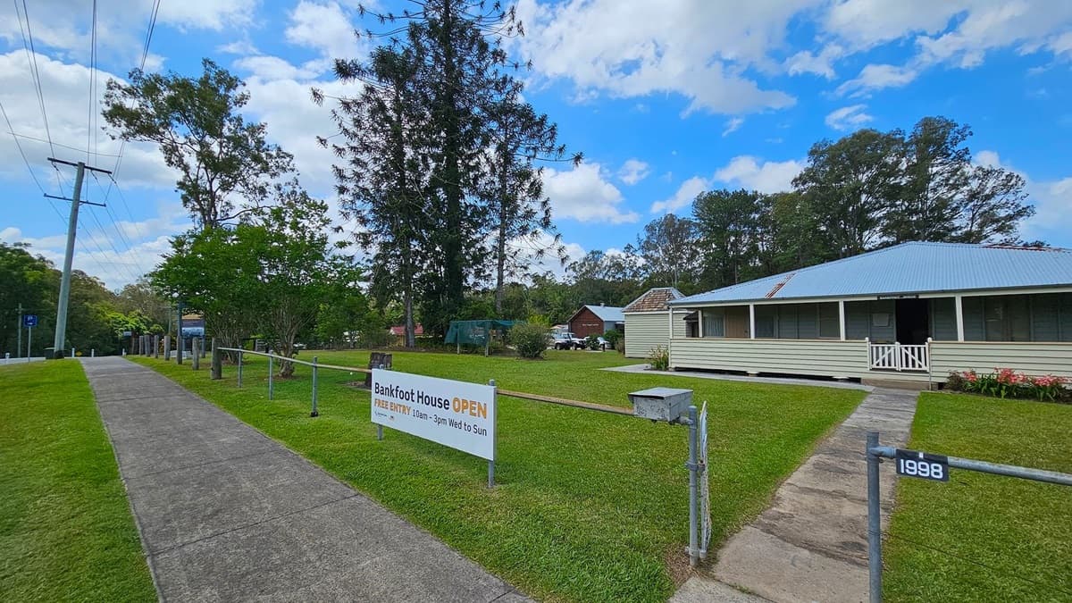 A paved footpath runs along the left side, bordered by grass and trees. A power line is visible above. On the right, a white wooden house with a blue roof and a small porch sits on a well-kept lawn. A sign in front reads "Bankfoot House OPEN" with visiting hours. A path leads from the gate to the house. In the background, more trees and a small building are visible.
