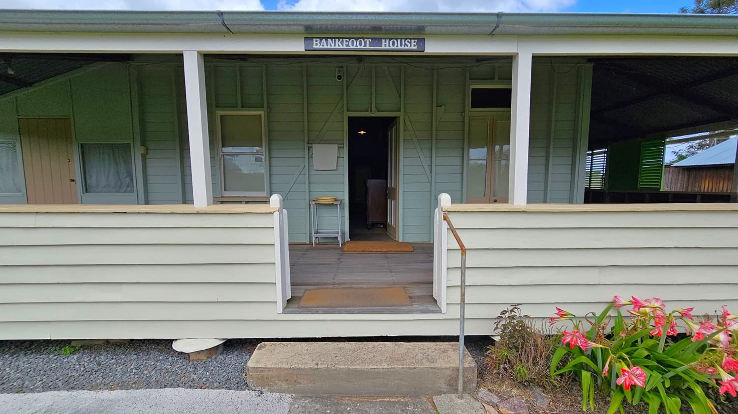 A wooden house with a sign reading "Bankfoot House" above the entrance. The exterior is painted in light green and grey. The entrance has a small porch with a wooden floor and a metal railing on the right. A doormat is placed at the doorway. On the right, there are pink and white flowers growing near the steps. The roof is metal, and the house has a simple, rustic appearance.