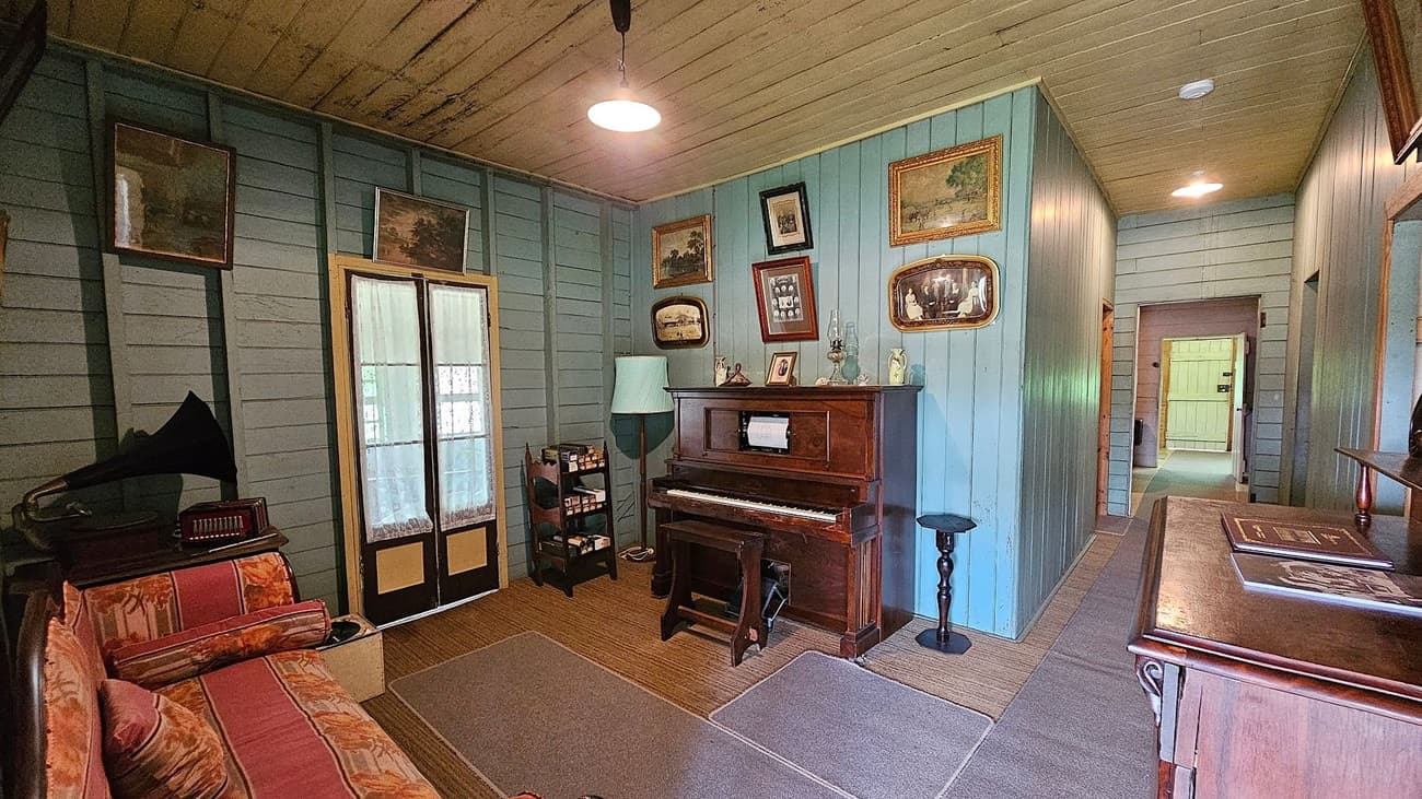A vintage room with wooden panelled walls painted in light blue. On the left, there's a patterned sofa with a gramophone on a side table. In the centre, a wooden piano is adorned with framed photos and a lamp. The right side features a hallway leading to another room. The floor is covered with woven rugs, and the ceiling is wooden with a hanging light fixture. Framed pictures decorate the walls.