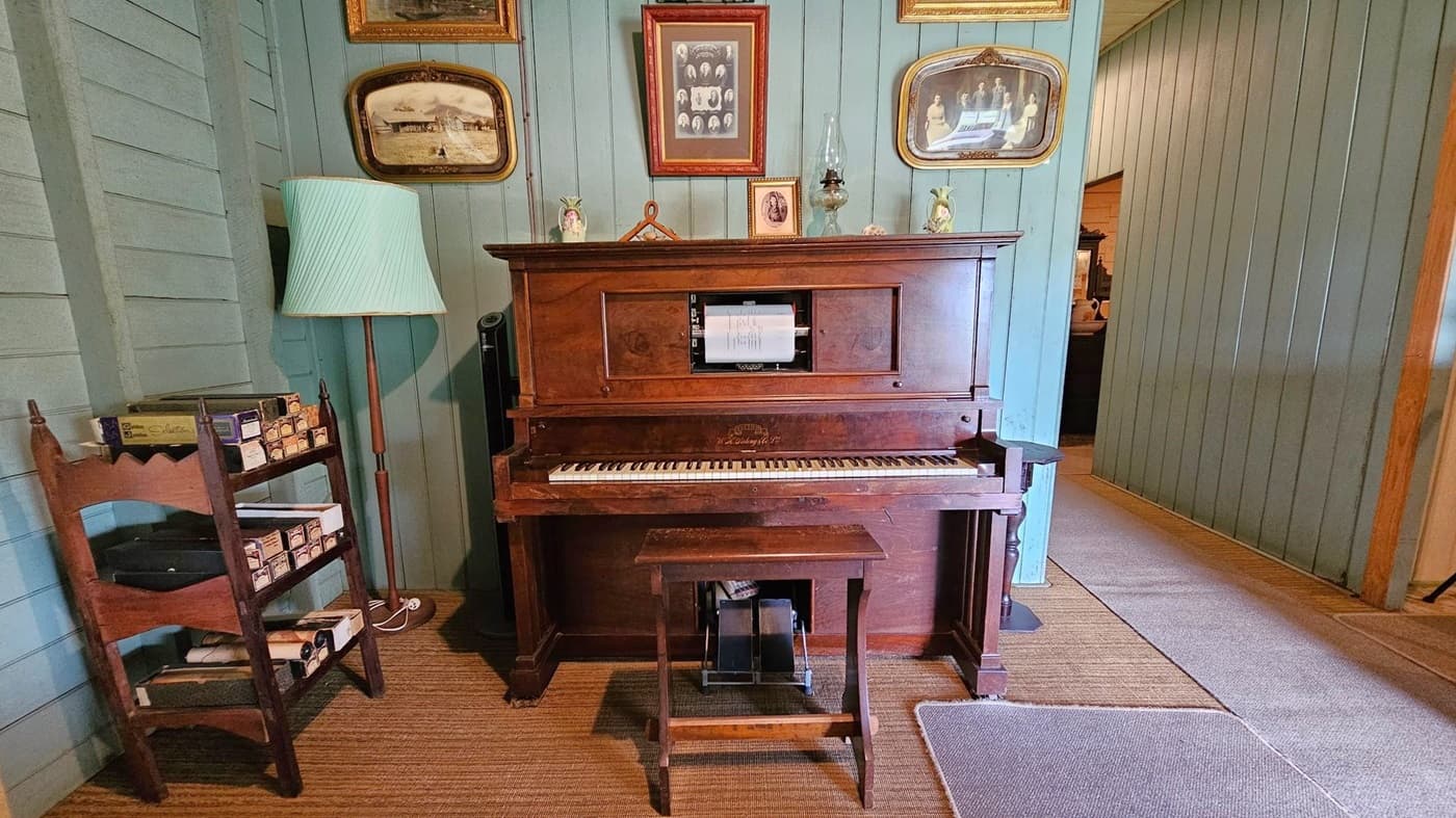 A vintage wooden piano stands against a light blue panelled wall. Above it are framed black-and-white photographs. To the left, a wooden shelf holds boxes and a tall lamp with a pleated shade. A small wooden stool is in front of the piano. The floor is covered with a woven rug and a grey mat. To the right, a hallway leads to another room.