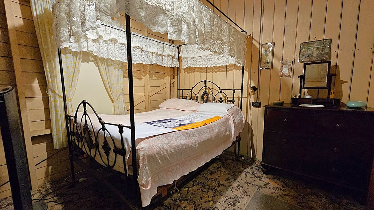 A vintage bedroom with a four-poster bed on the left, draped with lace curtains. The bed has a white coverlet and a decorative pillow. On the right, a dark wooden dresser holds a small mirror and some items. The walls are wooden panels with framed pictures hanging. The floor is covered with a patterned carpet.