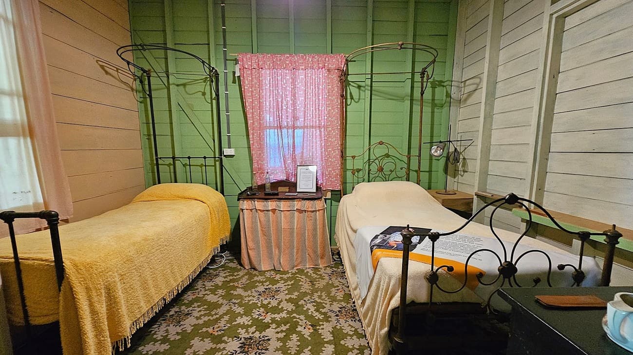 A room with two single metal-framed beds, each covered with a yellow blanket. The walls are painted green and beige. A small table with a pink skirt is positioned between the beds, holding framed documents. A pink curtain covers the window above the table. A vintage lamp is on the right side, and the floor is covered with a patterned carpet. A cup and a small item are on a black table in the foreground.
