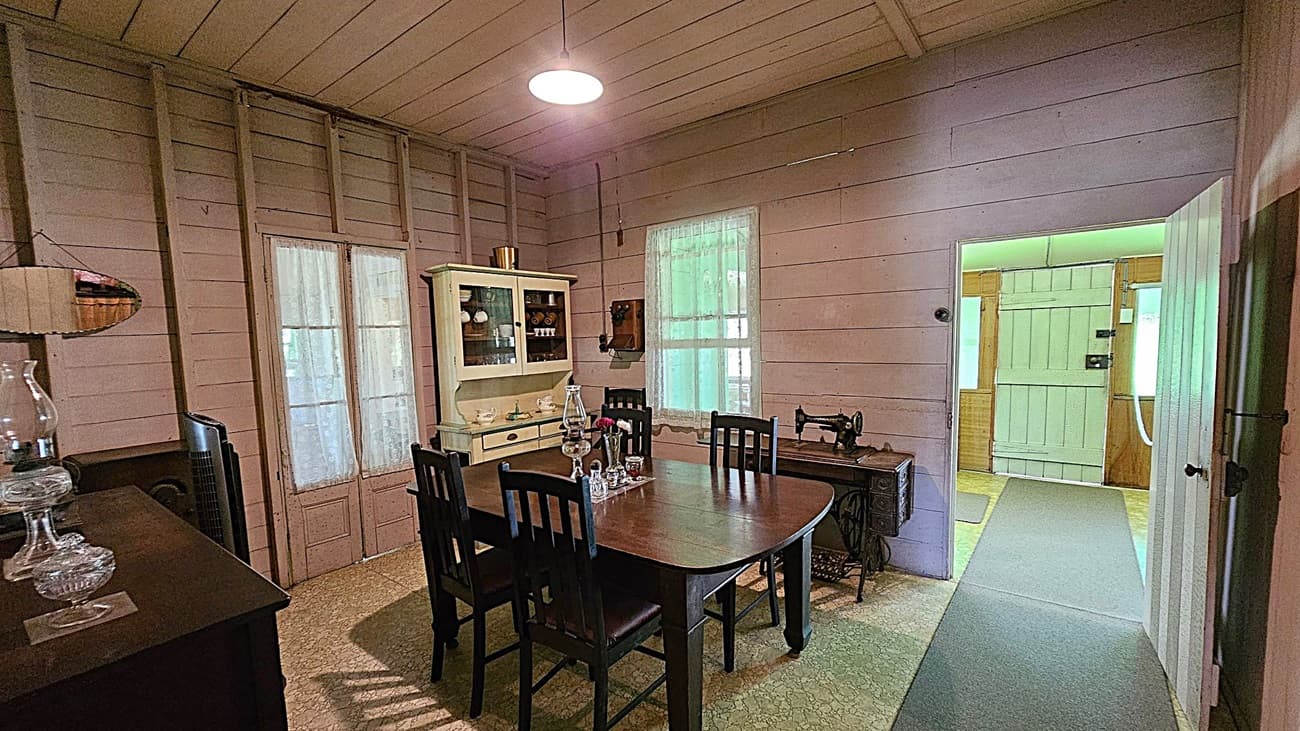 A vintage dining room with wooden walls and ceiling. On the left, a dark wooden sideboard holds glass lamps. In the centre, a wooden dining table with six chairs is set with glassware and a small vase. Behind the table, a cream-coloured cabinet displays crockery. To the right, an old sewing machine sits beneath a window. An open door leads to a hallway with greenish lighting. The floor is covered with patterned linoleum.