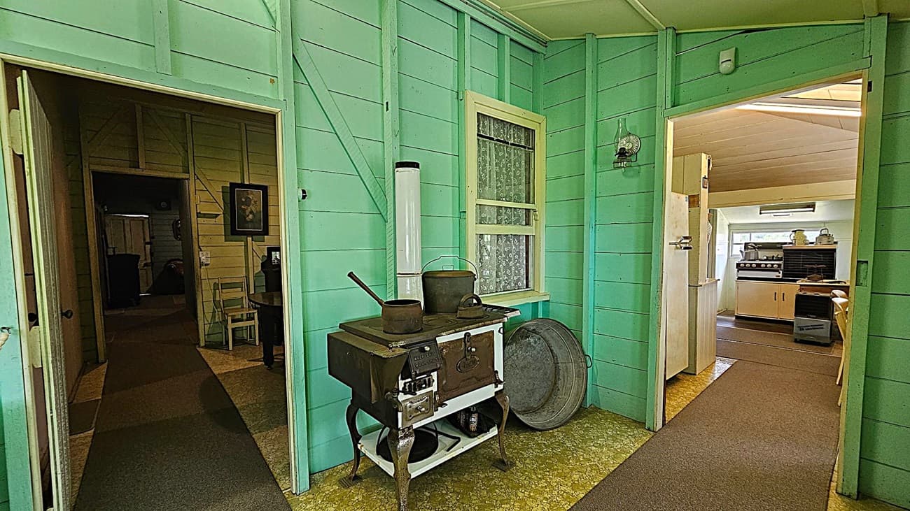 A room with light green wooden walls and a vintage stove in the centre. The stove has a pot and a kettle on top, with a large metal basin leaning against the wall. To the left, a doorway leads to a dimly lit room with a picture on the wall and a chair. To the right, another doorway opens into a kitchen area with a modern stove, cabinets, and a countertop. The floor is covered with a patterned linoleum, and the ceiling is plain and light-coloured.