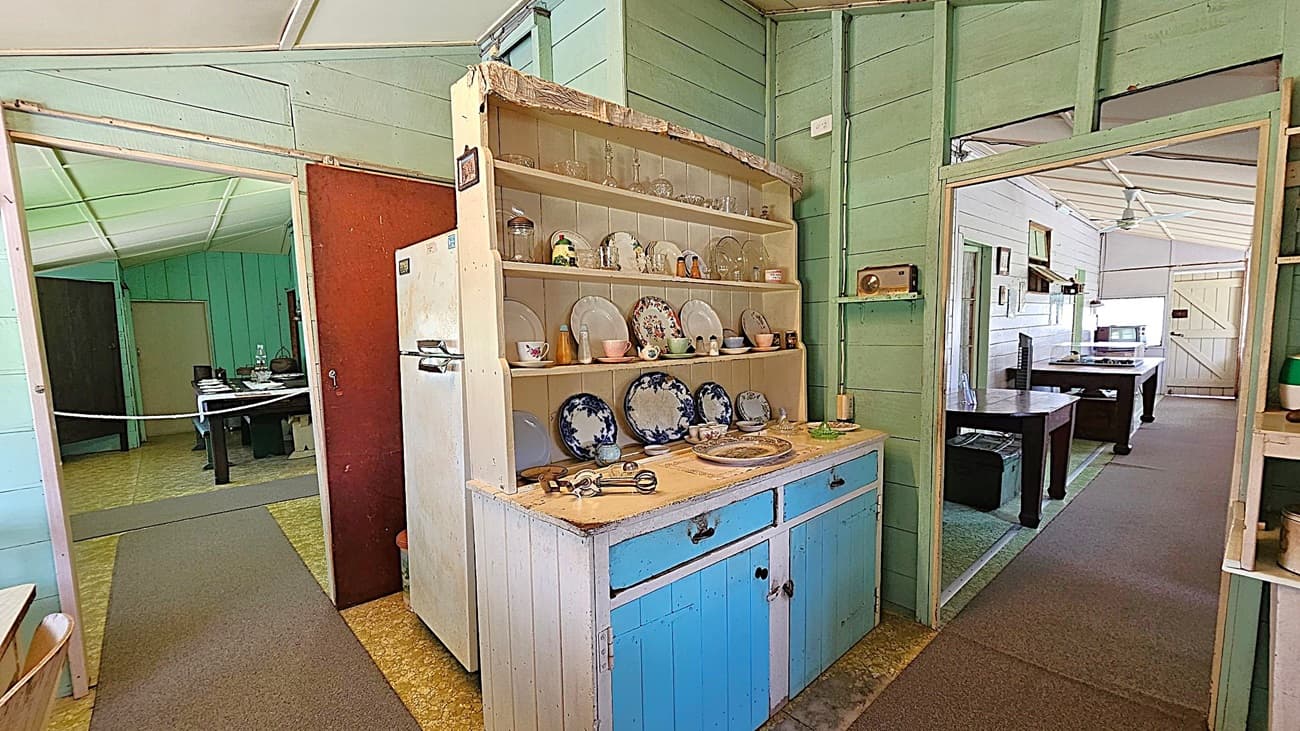 A vintage kitchen with light green wooden walls and a white ceiling. In the centre, a wooden cabinet with blue drawers displays various plates and cups. To the left, a doorway leads to another room with a table and chairs. On the right, another doorway reveals a room with a large table and a window. A white refrigerator stands next to the cabinet. The floor is covered with a patterned linoleum and a grey carpet runner. An old radio is mounted on the wall.