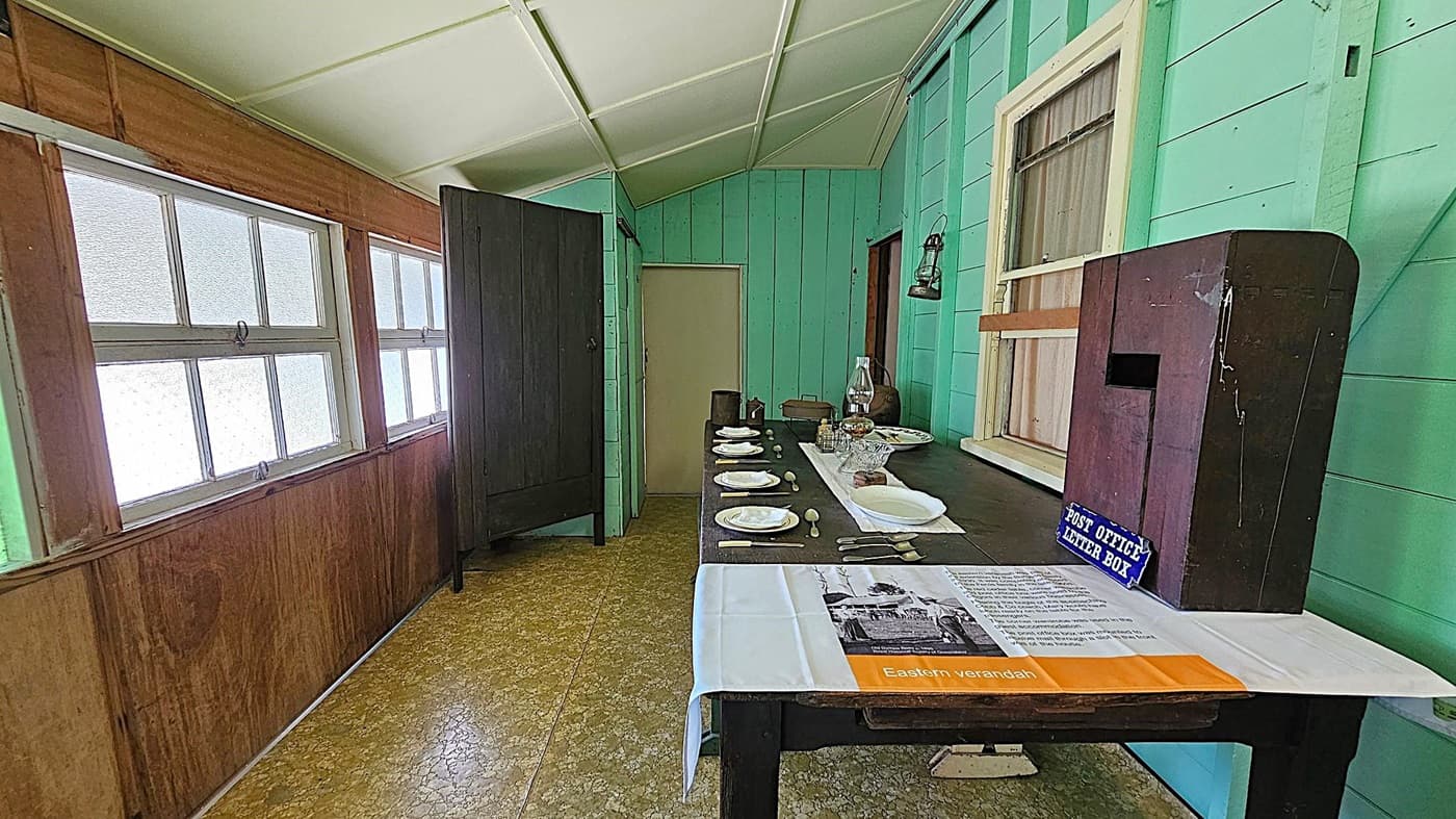 A room with light green wooden walls and a white ceiling. On the left, there are several frosted windows with wooden frames. In the centre, a long dark table is set with white plates, cutlery, and a glass jug. A lantern hangs on the wall above the table. On the right, a wooden post office letter box is placed on the table, with a sign and a paper describing "Eastern verandah". The floor is covered with a patterned linoleum.