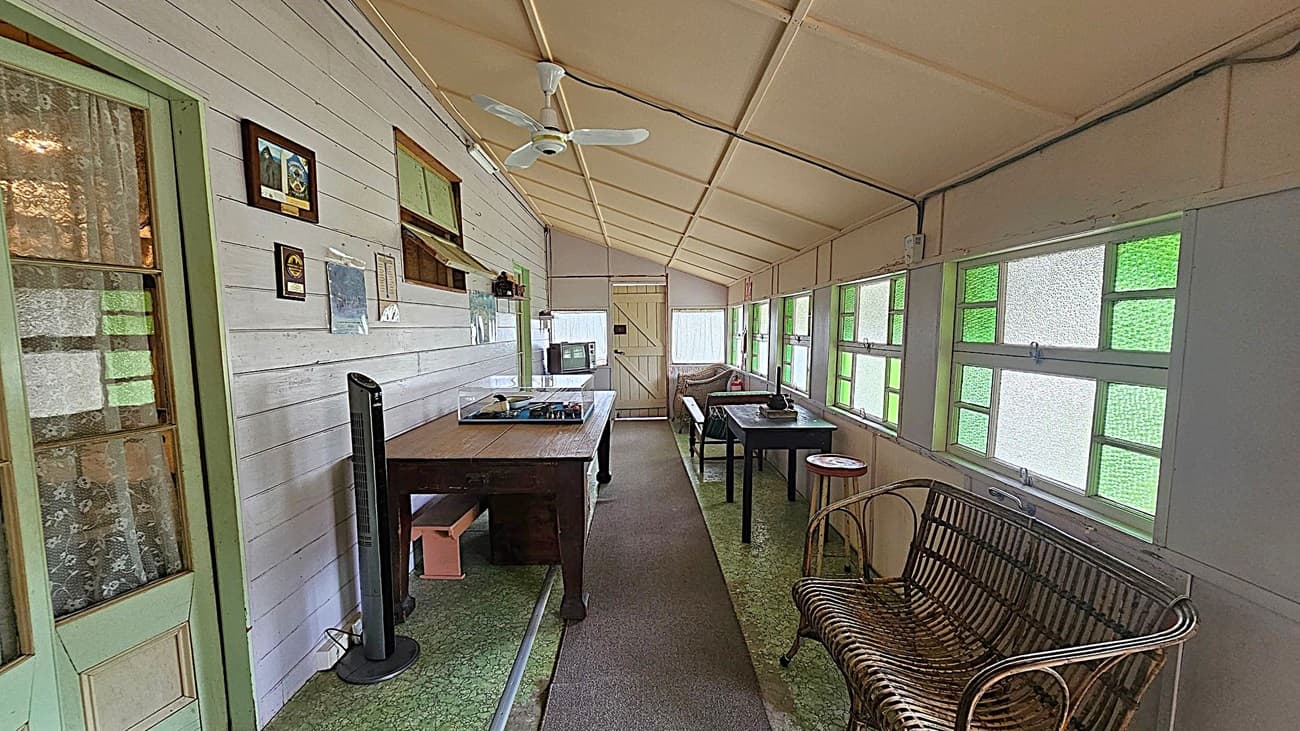 A narrow room with a slanted ceiling and green-tinted windows on the right. The left wall is white with framed pictures and a fan. A wooden table with a record player is in the centre, and a smaller table with a chair is further down. A wicker bench is near the windows. The floor is covered with a green patterned carpet and a grey runner. A door at the end of the room leads outside.