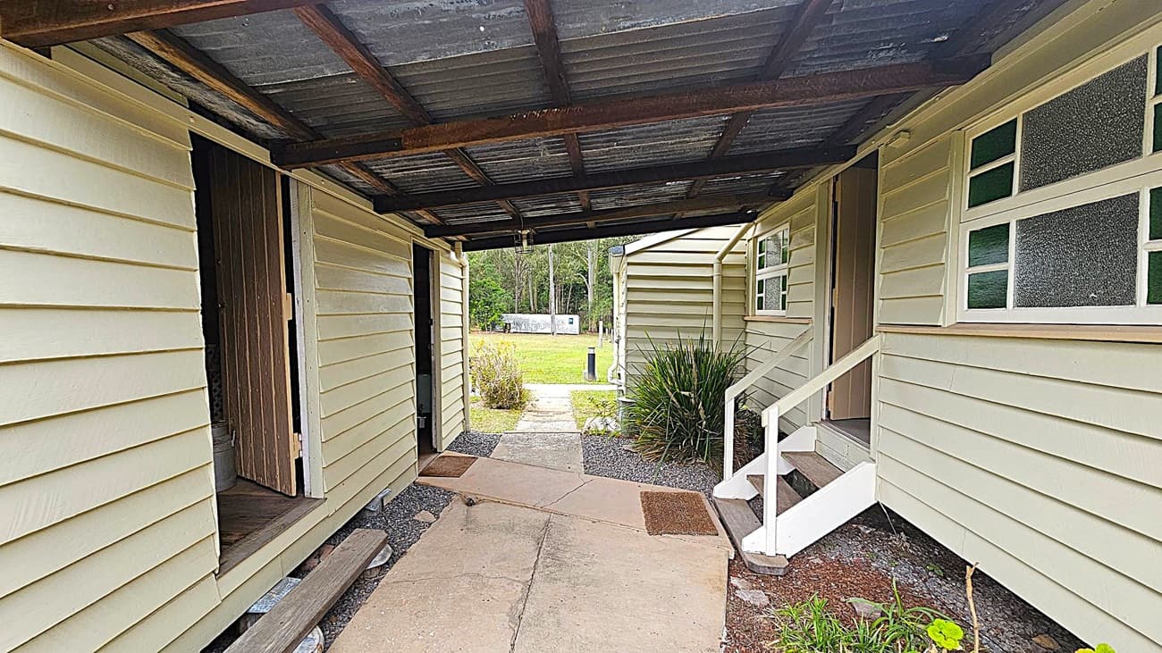 A covered walkway between two light green wooden buildings with corrugated metal roofing. On the left, there's an open door leading inside. On the right, a small set of white steps leads to another door. The ground is a mix of concrete and gravel, with a few doormats. In the background, there's a grassy area with bushes and trees, and a white structure is visible in the distance.
