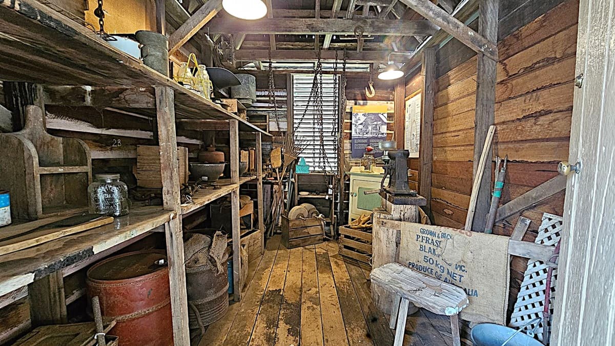 A rustic wooden shed interior with shelves on the left holding various tools, jars, and containers. The floor is made of wooden planks. In the centre, chains hang from the ceiling, and a window with slatted blinds is visible. On the right, a small table with a grinder, a wooden chair, and a sack are present. The walls are made of wooden planks, and the ceiling has exposed beams with hanging lights. Various gardening tools lean against the right wall.