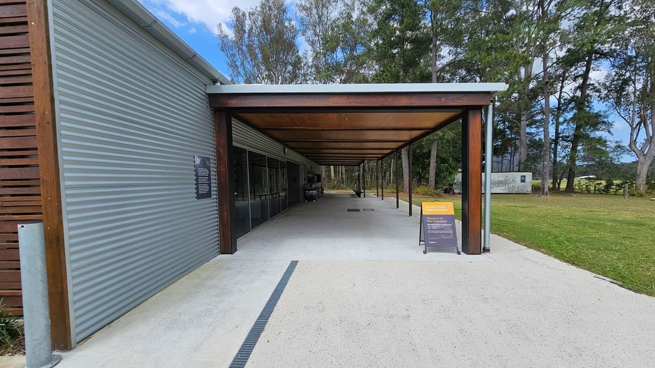 A building with a wooden facade labelled "Mary Grigor Centre" is in the centre. To the left, there's a large metal water tank and a hedge with trees behind it. The foreground shows a parking area with a blue disabled parking sign and yellow markings. On the right, a covered area extends from the building, with a signboard near the entrance. Tall trees are in the background, surrounding the area.