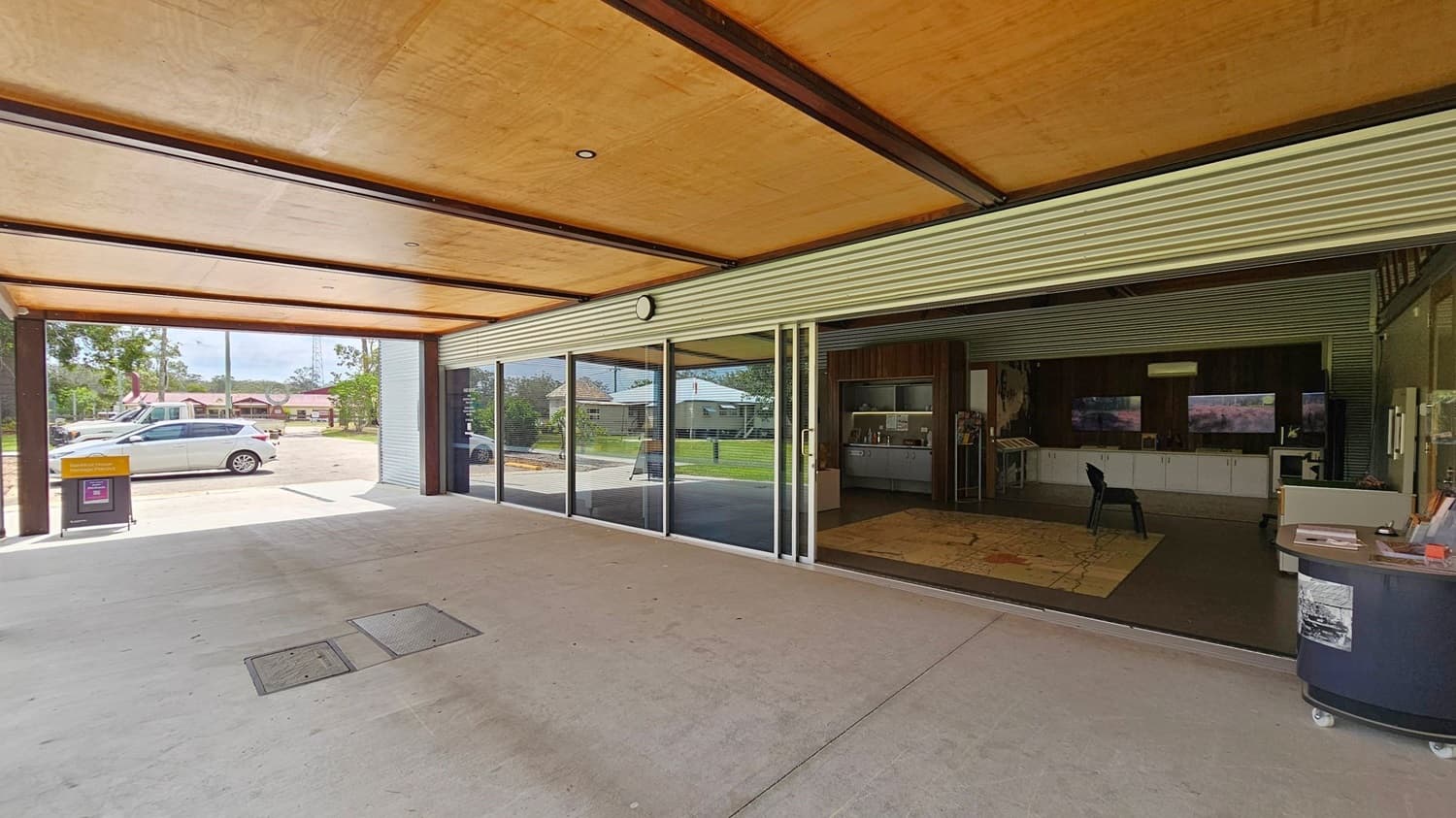 A covered outdoor area with a wooden ceiling and concrete floor. On the right, there is a building with large glass sliding doors, revealing an interior with a chair, cabinets, and a display area. On the left, outside, there is a white car parked on a paved area, with a small bin nearby. In the background, there are trees and a few houses.