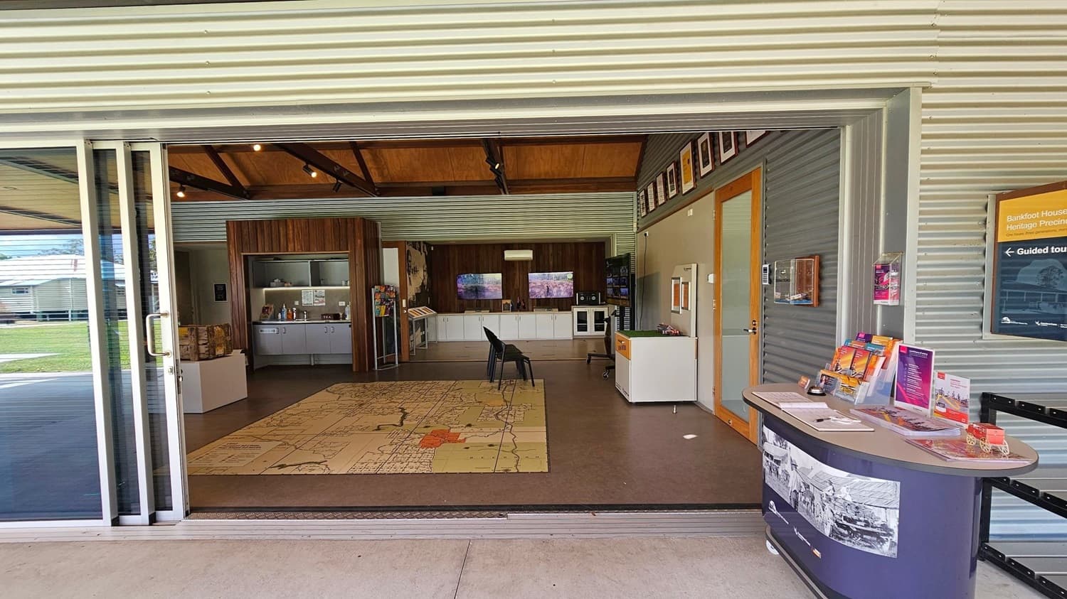A room with corrugated metal walls and a high wooden ceiling. In the foreground, a table displays brochures and pamphlets. On the right wall, a sign indicates guided tours. The floor features a large map design. In the background, there are cabinets, a display area, and two black chairs. The room is well-lit with natural light from the open sliding glass doors. Outside, a grassy area is visible.