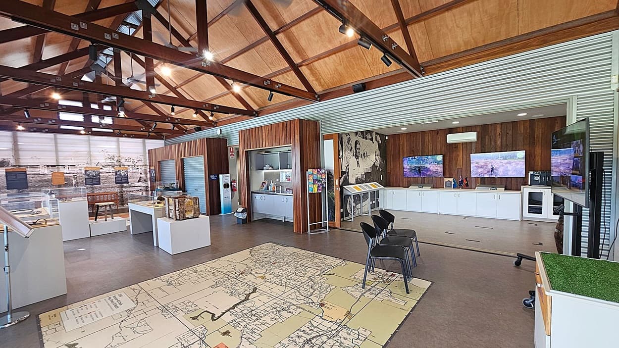 A spacious room with a high wooden ceiling and exposed beams. On the left, there are display cases with artefacts and a large historical photo on the wall. In the centre, a large map is printed on the floor, with a few chairs nearby. On the right, there are three screens mounted on a wooden panelled wall, above white cabinets. A small kitchenette area is visible, along with a brochure stand. The floor is concrete, and the room is well-lit with spotlights.