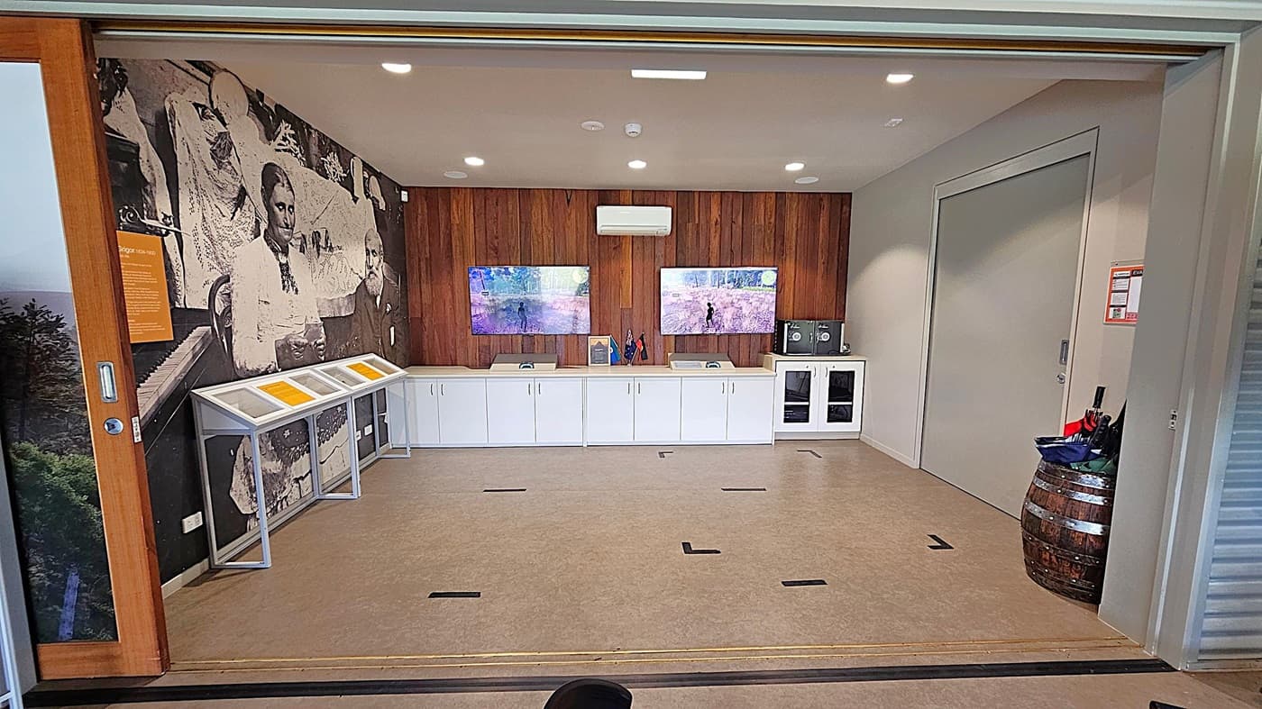 A room with a large mural on the left wall depicting historical figures. Below the mural are display cases with informational panels. The back wall is wooden with two screens showing outdoor scenes. Below the screens are white cabinets with small flags and items on top. On the right, there's a closed door and a wooden barrel holding umbrellas. The floor is carpeted, and the ceiling has recessed lighting.