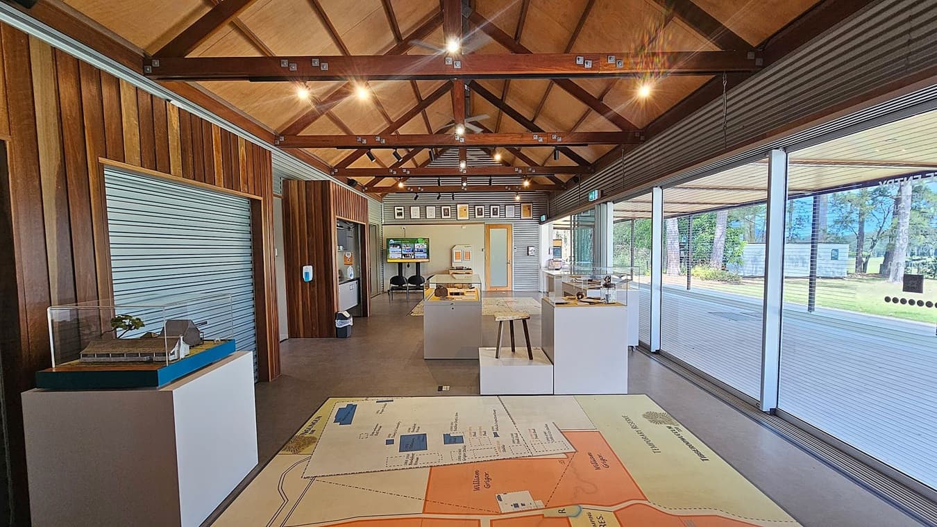 Interior of a modern exhibition space with a high, wooden-beamed ceiling. On the left, a model of a building is displayed on a pedestal. The right side features large glass windows offering a view of trees and grass outside. In the centre, there are display cases and a large map on the floor. The back wall has framed pictures and a digital screen. The floor is smooth and grey, and the walls are a mix of wood and metal panelling.