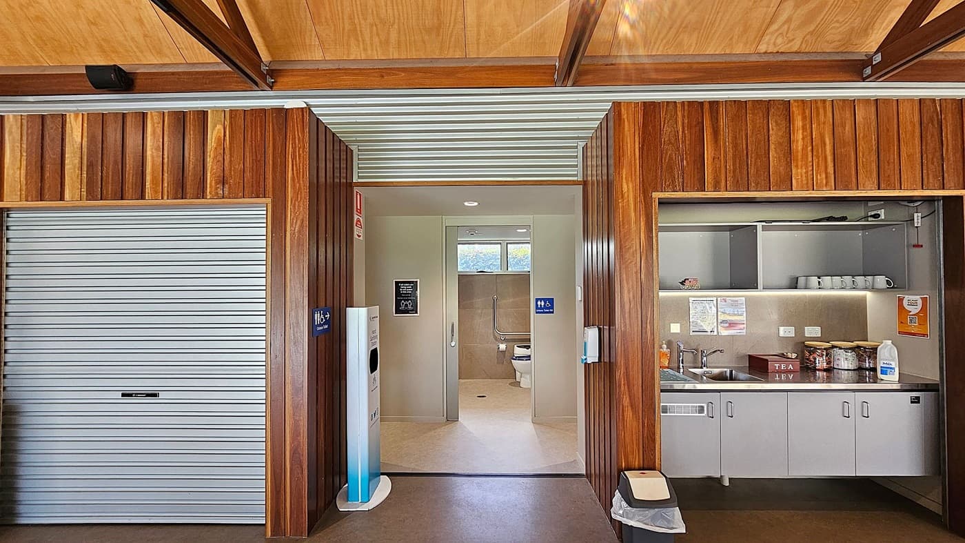 A room with wooden walls and a wooden ceiling. On the left, there's a closed metal roller door. In the centre, an open doorway leads to a bathroom with a toilet and grab bars visible. A hand sanitiser dispenser is mounted on the wall near the entrance. On the right, there's a kitchenette with a sink, mugs, and various containers on the counter. Open shelves above hold additional items. A small bin is on the floor near the kitchenette.