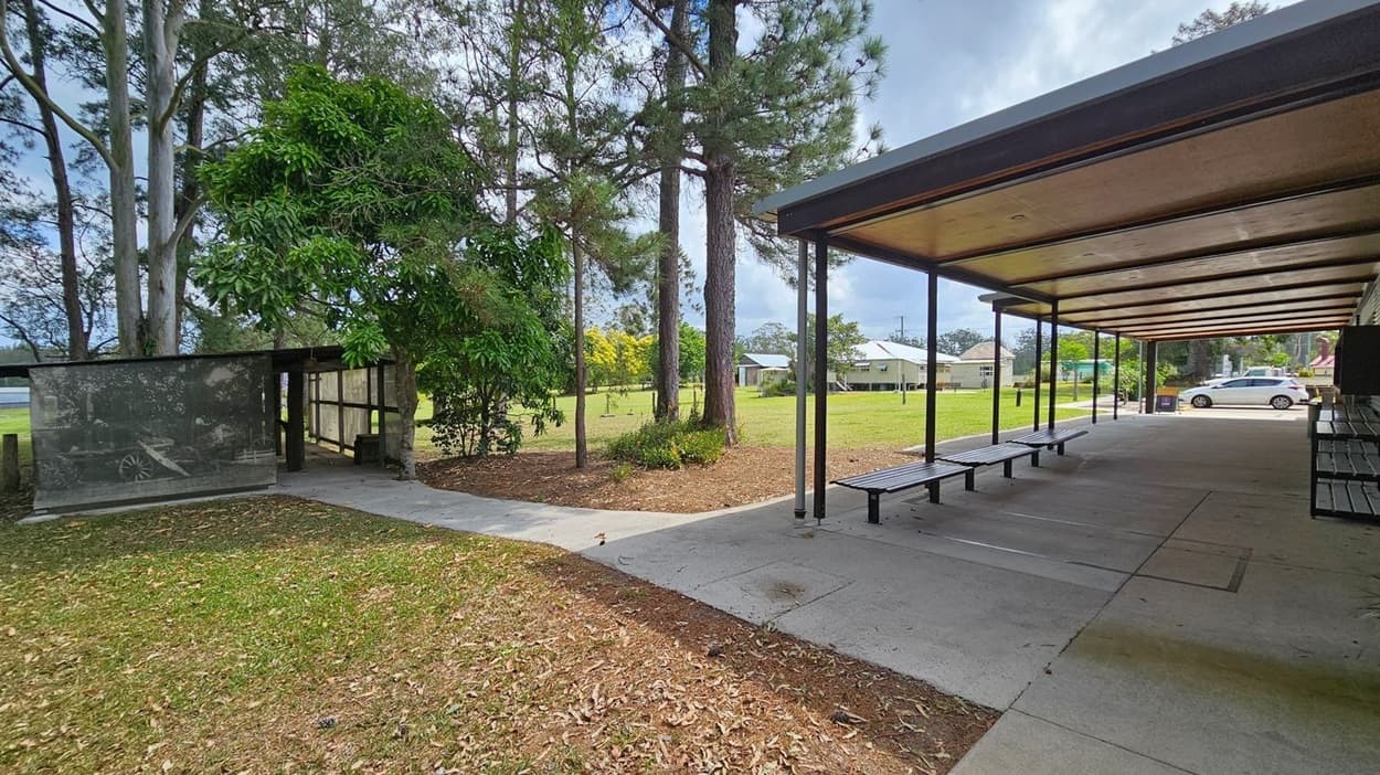 A covered walkway with a wooden ceiling and metal supports is on the right, featuring benches along its length. To the left, a path leads past a mural-covered wall and several large trees, including a lush green one. In the background, a grassy area with more trees and a few houses is visible. A white car is parked near the houses. The ground is a mix of concrete, grass, and scattered leaves.