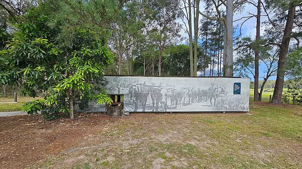 A mural on a long, rectangular wall features an image of people and oxen. On the left, dense green foliage partially obscures the mural. The background is filled with tall trees, and the ground is covered with grass and scattered leaves. A small sign is visible on the right side of the mural.