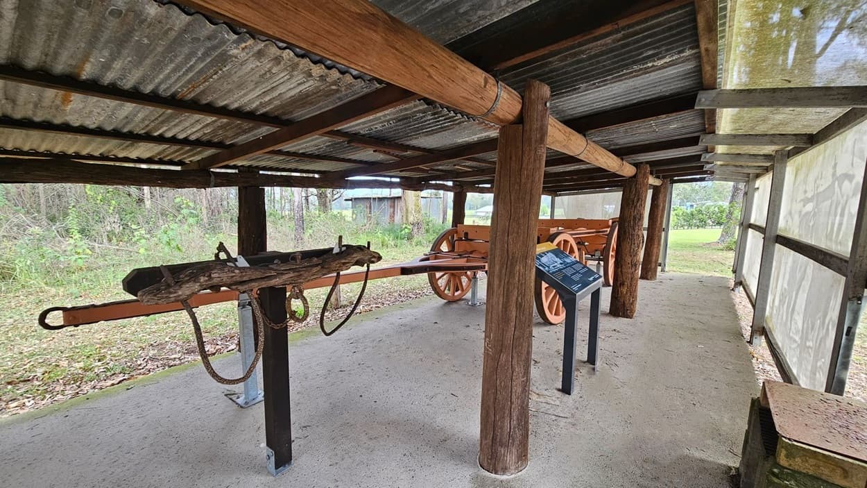 A rustic shelter with a corrugated metal roof supported by wooden beams. Inside, there is a wooden cart with large orange wheels on the right. A wooden yoke with metal fittings is mounted on a stand to the left. An informational sign is positioned near the cart. The floor is concrete with scattered leaves. The background shows a grassy area and trees.