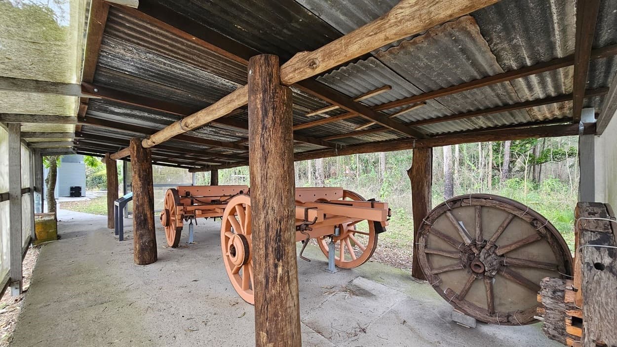 A rustic shelter with a corrugated metal roof supported by wooden beams. Underneath, there are two large wooden carts with spoked wheels, painted in a light brown colour. To the right, a large wooden wheel leans against the wall. The ground is covered with concrete, and there are scattered leaves. In the background, there is a grassy area with trees.