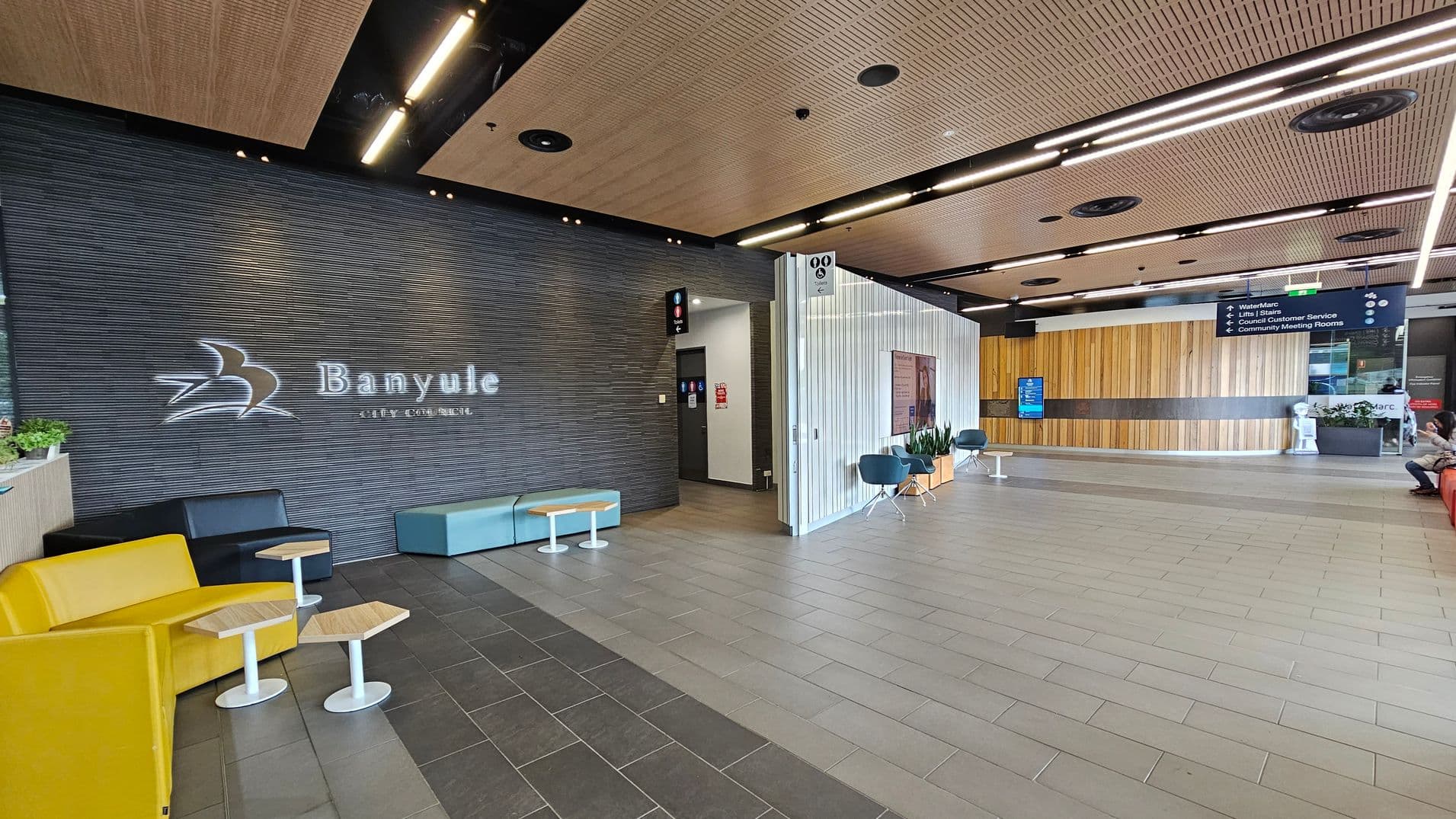 A modern interior space with a dark textured wall on the left displaying the words "Banyule City Council" and a logo. Below are yellow and black seating with small wooden tables. The floor is tiled in grey. The ceiling features wooden panels with recessed lighting. On the right, a light-coloured wall with signage and blue chairs. A wooden panelled wall is in the background, with a digital screen and a person seated on a red bench.