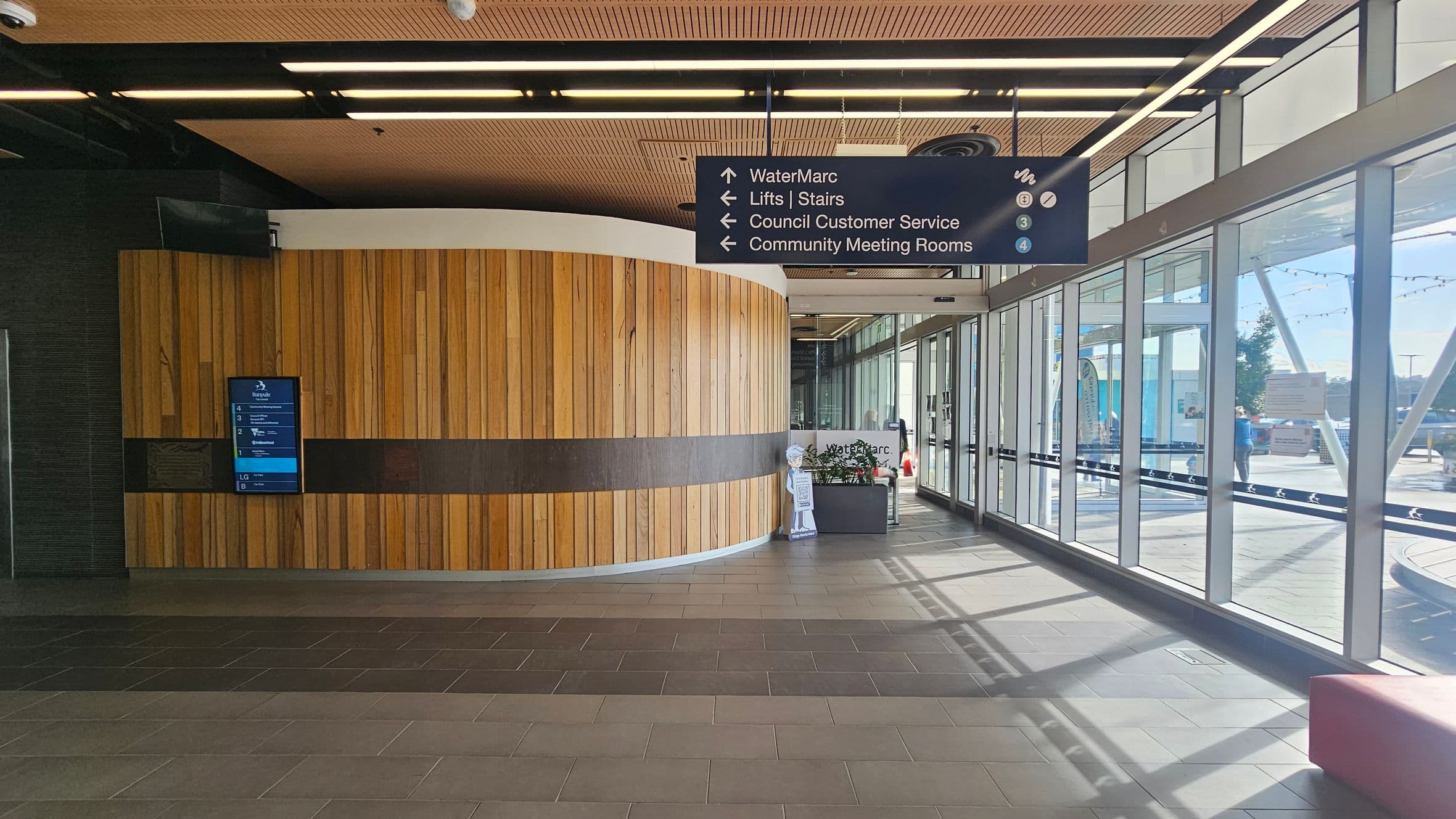 A modern interior with a curved wooden wall on the left, featuring a digital directory. Above, a sign directs to WaterMarc, lifts, stairs, council customer service, and community meeting rooms. On the right, large glass windows allow natural light, with a view of the outside. The floor is tiled in shades of grey, and the ceiling has wooden panels with strip lighting. A small reception area is visible near the windows.