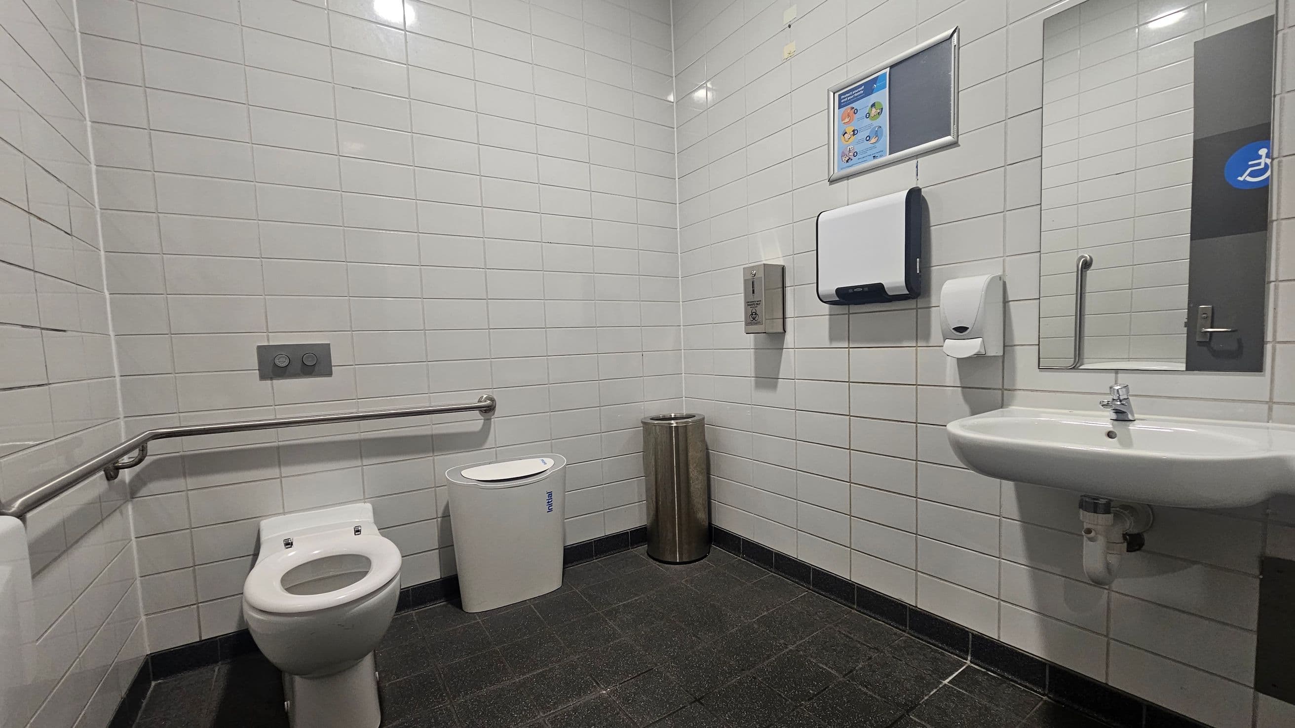 A public restroom with white tiled walls and a dark tiled floor. On the left, there is a toilet with a metal grab bar beside it. Next to the toilet is a white sanitary bin and a metallic waste bin. On the right, a wall-mounted sink with a soap dispenser above it. Above the sink, a mirror and a paper towel dispenser are visible. A sign with instructions is mounted on the wall. The ceiling has recessed lighting.