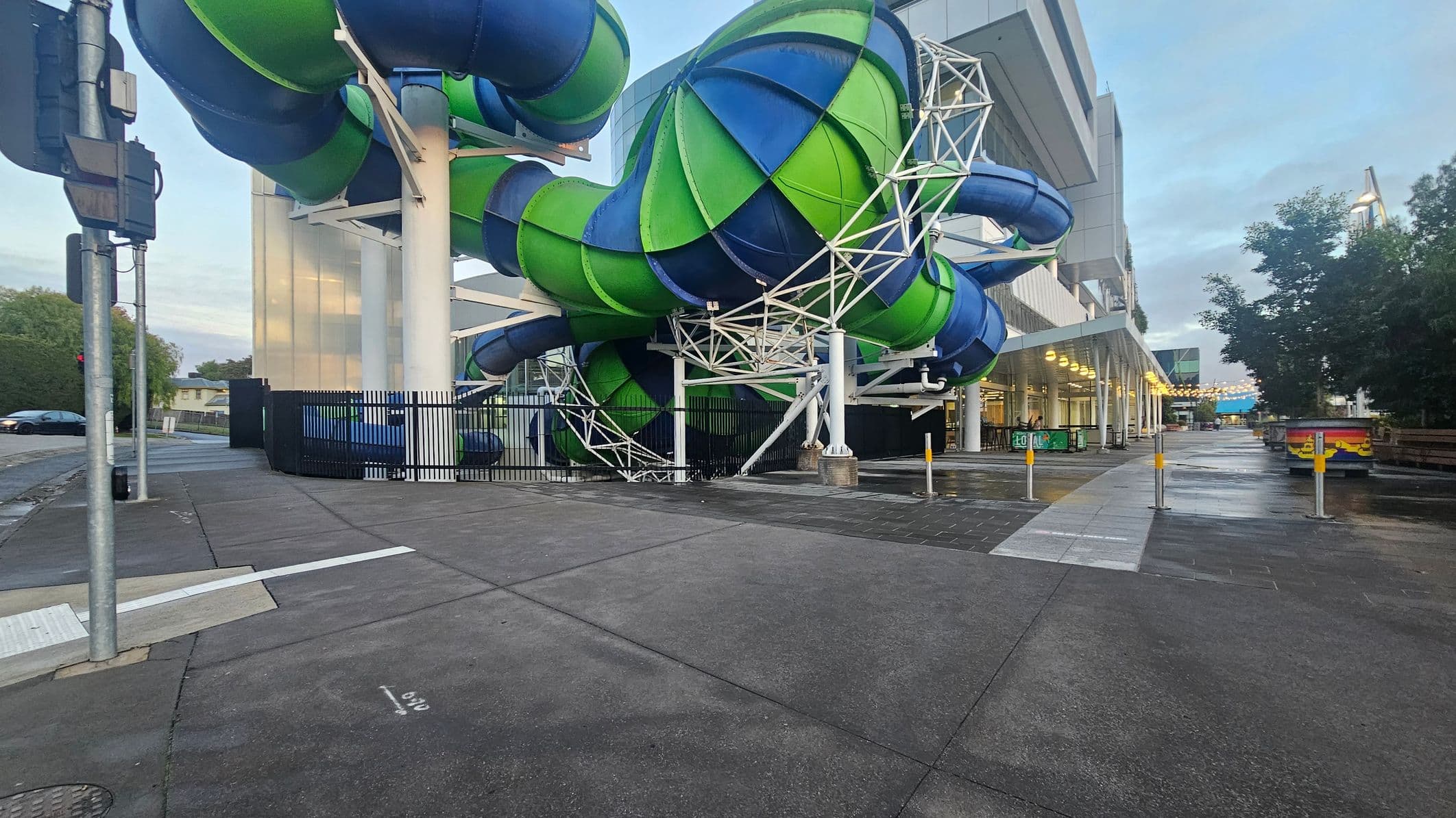 Large, intertwined water slides in green and blue dominate the centre, supported by white metal structures. On the right, a modern building with a covered walkway is lit by rows of lights. The ground is a mix of concrete and wet pavement, with bollards lining the path. On the left, a traffic light and a parked car are visible. Trees and greenery are in the background, adding contrast to the urban setting.