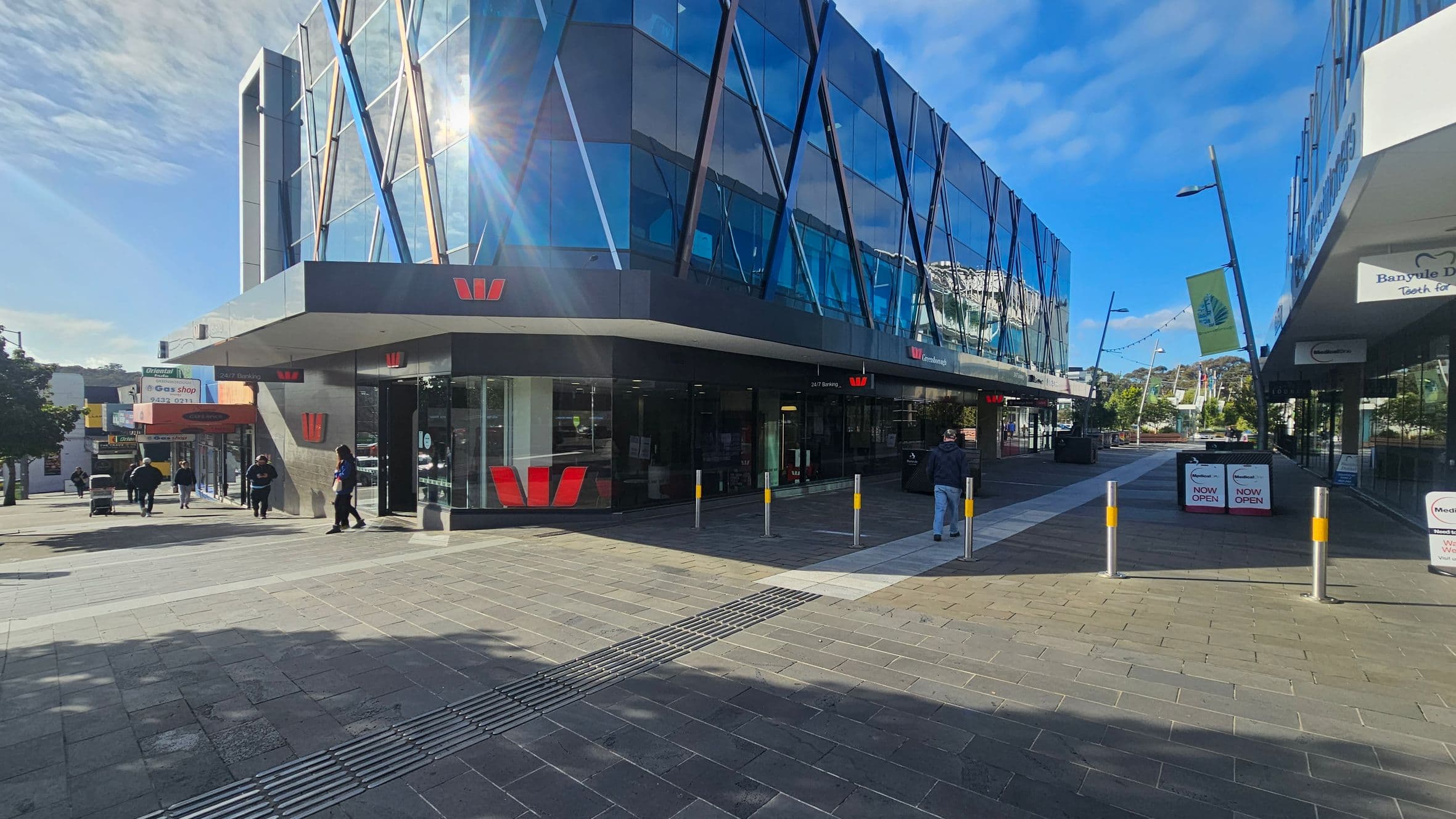 A modern building with a glass facade featuring diagonal blue and white lines is on the left. The building has a prominent red logo. People are walking on the pavement in front of it. On the right, there are more shops with signs and a pathway leading further into the distance. The ground is paved with grey and reddish tiles, and there are several bollards near the entrance. The sky is clear and blue.