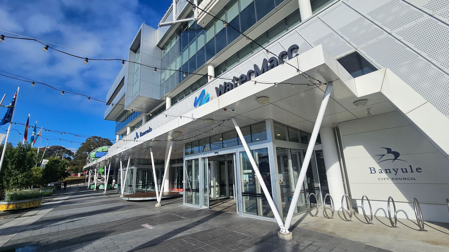 A modern building with a glass facade and angular design elements. The entrance features large glass doors and a sign reading "WaterMarc" above. To the right, there's a "Banyule City Council" sign. Several white poles support the structure, and string lights are suspended above. Flags are visible on the left, and there are trees and planters along the paved walkway. The sky is clear and blue.