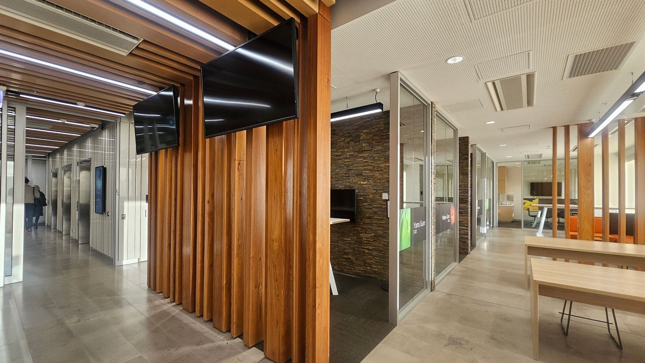 A modern office interior with a corridor on the left featuring elevators and a person walking away. The ceiling has wooden slats with integrated lighting. On the right, there are glass-walled meeting rooms with stone-textured walls and wooden partitions. A wooden table with chairs is in the foreground. Two black screens are mounted on the wooden slats. The floor is tiled, and the ceiling has recessed lighting and ventilation panels.