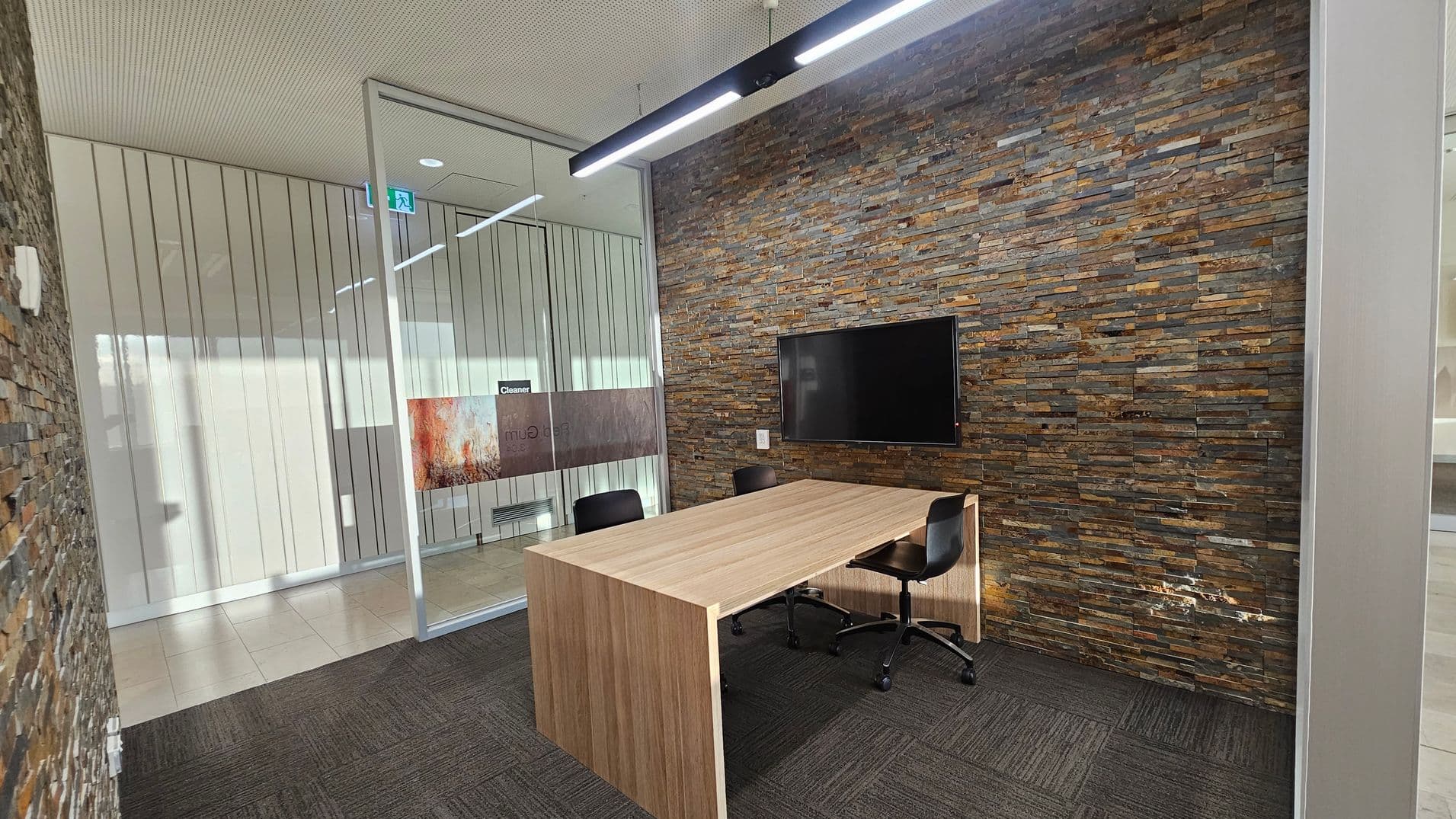 A modern office space with a textured stone wall on the right, featuring a mounted flat-screen TV. In the centre, a wooden table with two black swivel chairs. The left side has a glass partition with vertical lines, leading to another room. The floor is covered with dark grey carpet tiles, and the ceiling is light-coloured with a linear light fixture.