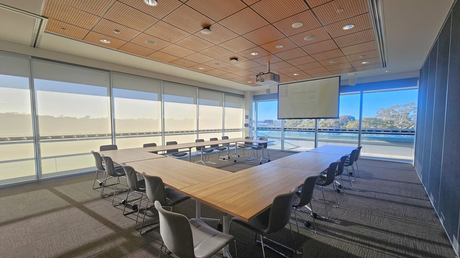 A conference room with a U-shaped arrangement of wooden tables surrounded by grey chairs. The left side features large windows with blinds partially drawn, allowing natural light to enter. On the right, a projector screen hangs from the ceiling, displaying faint text. The background shows a view of trees through the windows. The ceiling is panelled with wood and fitted with recessed lighting. The floor is carpeted in a dark tone.