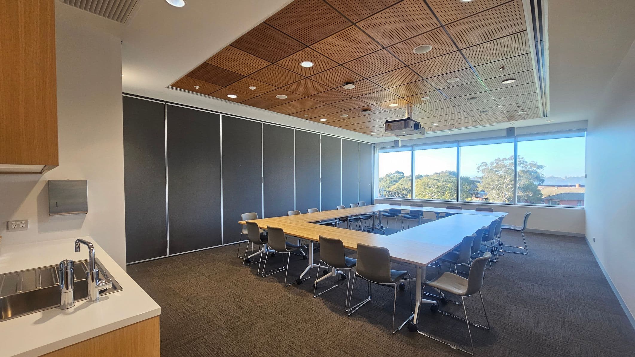 A conference room with a U-shaped arrangement of wooden tables and grey chairs on a carpeted floor. On the left, there's a kitchenette with a sink and a paper towel dispenser. The back wall features large windows with a view of trees and rooftops. The ceiling is wooden with recessed lighting and a projector.