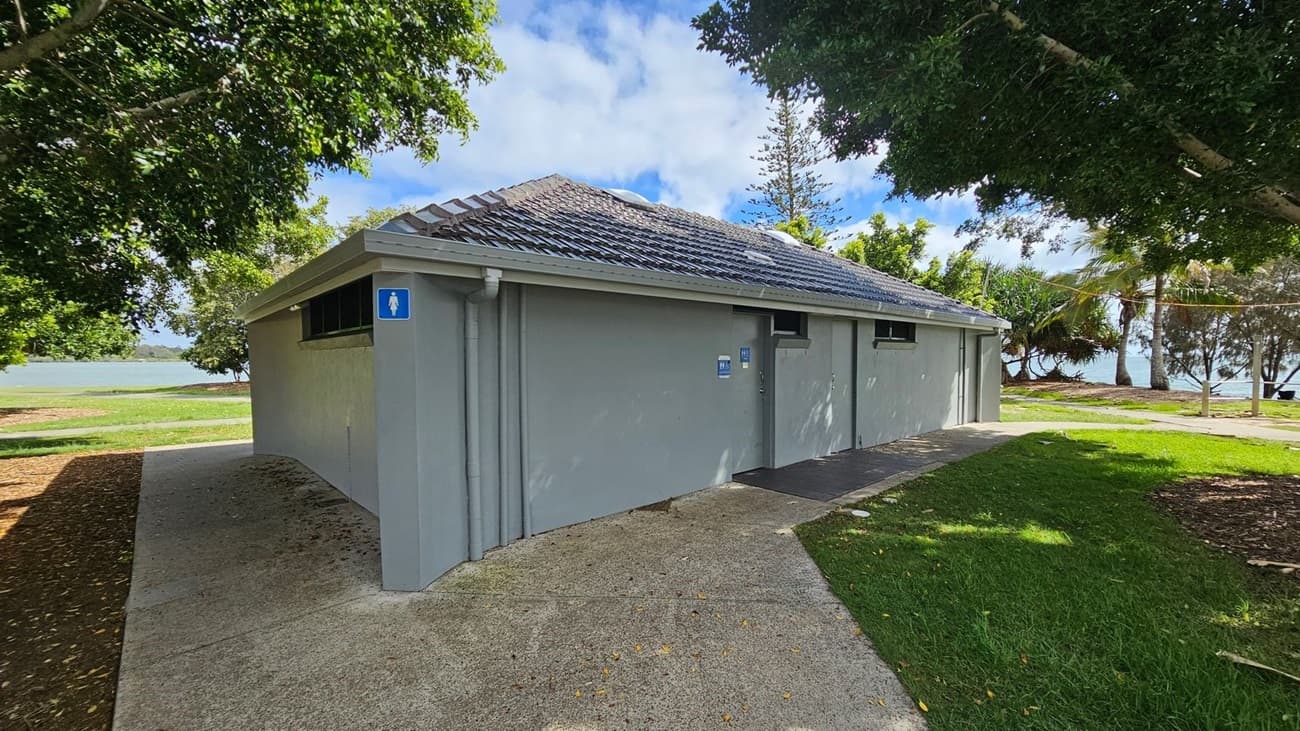 A grey public restroom building with a tiled roof is surrounded by trees and grass. On the left, a path leads towards a body of water visible in the background. The building has signs indicating male and accessible facilities. The ground is a mix of concrete and grass, with some fallen leaves. Trees provide shade, and there are glimpses of the water and more trees in the distance.