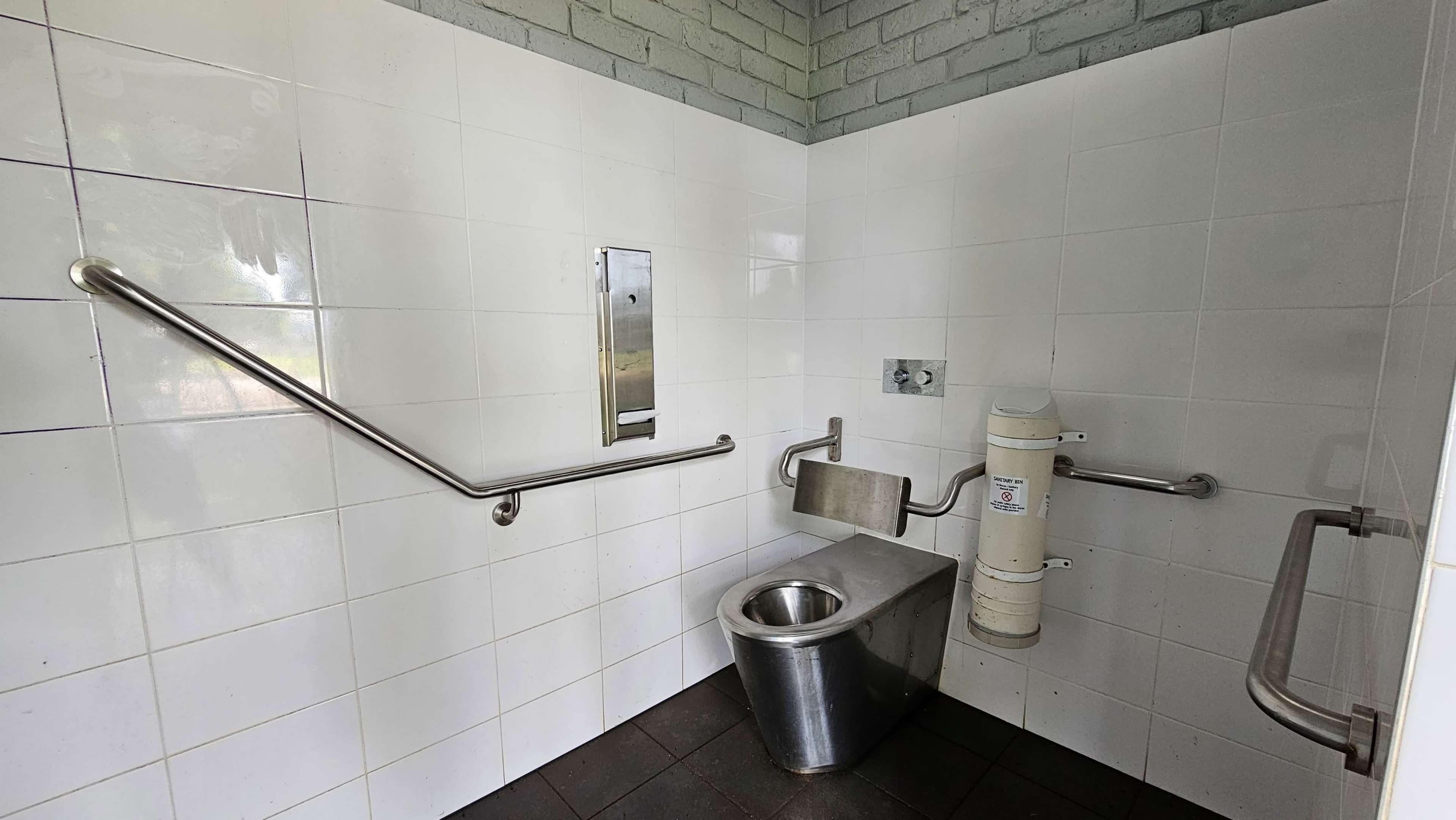 A public toilet with white tiled walls and a dark tiled floor. In the centre, there is a stainless steel toilet with a backrest. On the right, a wall-mounted baby changing station is visible. Stainless steel grab bars are installed on the left and right walls for accessibility. The upper part of the walls is made of light-coloured bricks.