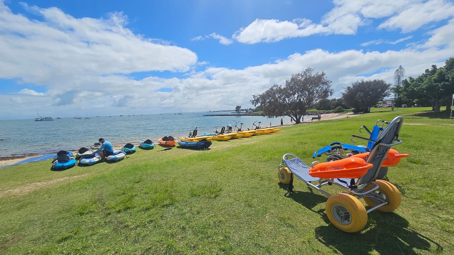 On the left, a beach wheelchair with large yellow wheels and orange armrests sits on grass. On the right, another beach wheelchair with grey wheels is nearby. In the middle, several kayaks and yellow water bikes are lined up on the grass near the shoreline. The background features a calm sea with a few boats and a rocky breakwater. Trees are visible on both sides.