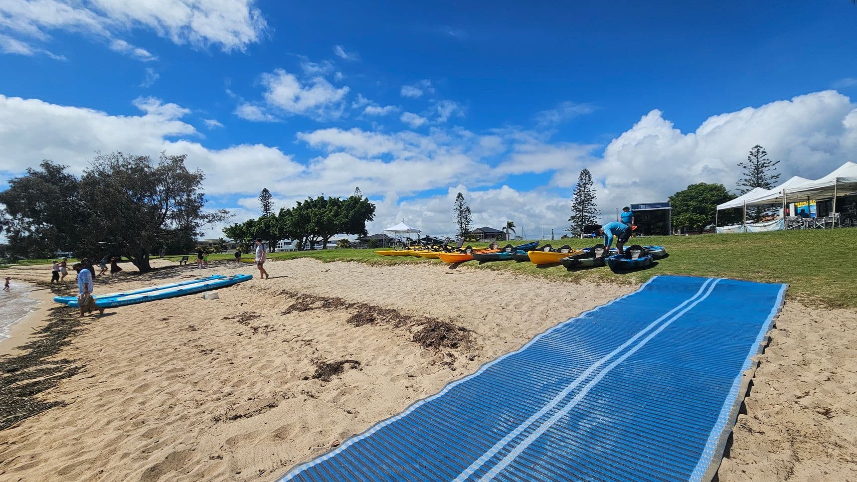 A sandy beach with a blue mat leading from the foreground to the water. On the left, people are near the shoreline with paddleboards. In the middle, kayaks are lined up on the grass. On the right, a person is handling equipment near a row of white tents. Trees and a few buildings are visible in the background.