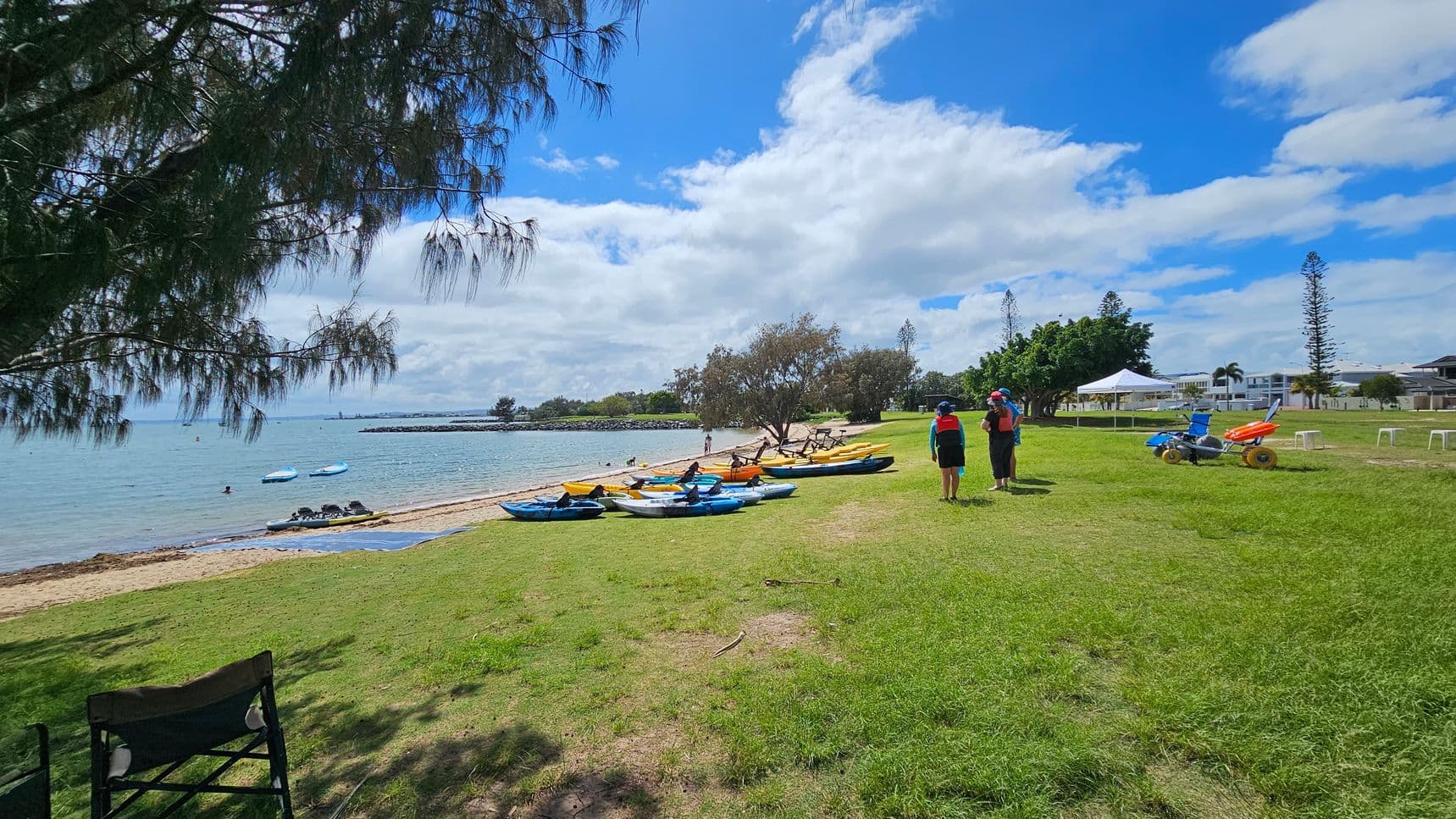 A beach scene with a grassy area in the foreground. On the left, a tree with overhanging branches provides shade. Several kayaks and paddleboards are lined up on the sand near the water. Two people wearing life jackets stand on the grass, facing the water. In the background, there are trees, a white tent, and some buildings. A beach wheelchair is parked on the grass to the right. The water is calm, with a few people swimming.