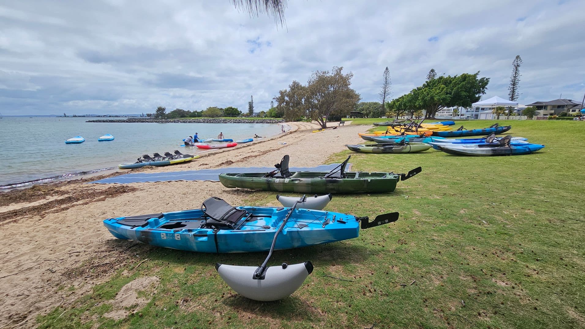 Kayaks in various colours, including blue, green, and yellow, are lined up on a grassy area to the right. The sandy beach stretches along the middle, leading to a calm body of water on the left. Several people are near the water's edge, with a few kayaks floating nearby. Trees and a few buildings are visible in the background, providing a scenic backdrop.