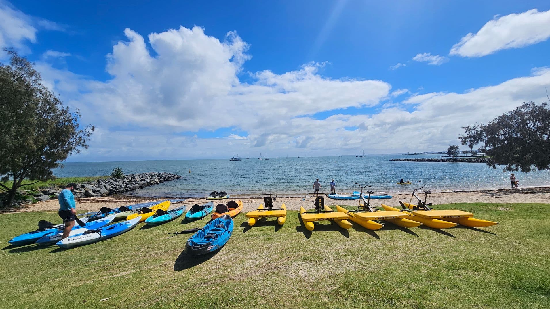 On the left, a person in a blue shirt stands near several blue and yellow kayaks on a grassy area. In the middle, more kayaks and yellow water bikes are lined up on the grass, with the beach and ocean in the background. Two people are near the water's edge, and a few boats are visible on the horizon. On the right, a person sits under a tree, with more trees framing the scene. The sky is partly cloudy.