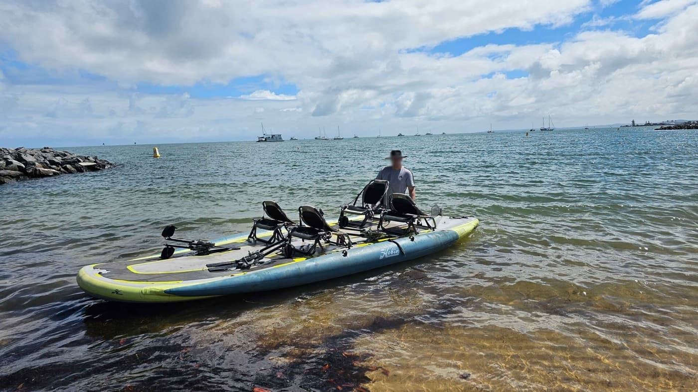 A man wearing a hat stands on a large paddleboard with multiple seats, positioned in shallow water near the shore. The water is clear, revealing a sandy and rocky bottom. To the left, a rocky breakwater extends into the sea. In the background, several boats and yachts are anchored in the distance, with a buoy visible in the water. The sky is partly cloudy.