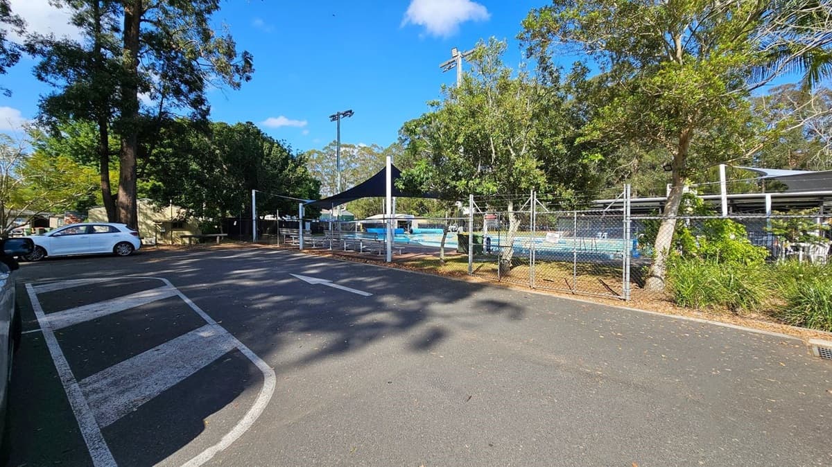 A car park with a white car on the left. The road has white painted lines and an arrow. On the right, there's a fenced area with a swimming pool covered by a black shade sail. Trees and greenery surround the area, with tall trees in the background. The ground is paved, and there are shadows from the trees.