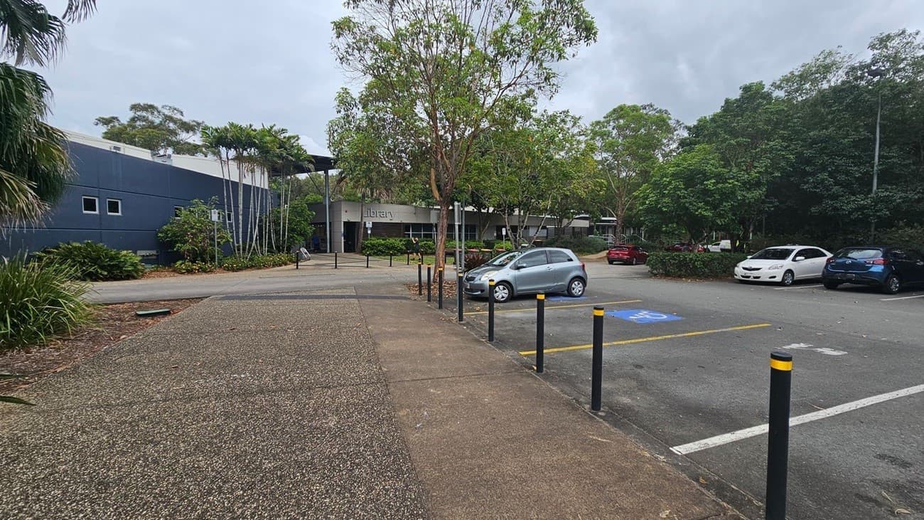 A paved walkway leads to a building labelled "library" in the centre. On the left, there are tall palm trees and a dark grey building with small windows. On the right, a car park with several vehicles, including a silver car in a disabled parking space. The area is surrounded by lush green trees and foliage. Black bollards with yellow tops line the walkway and car park.