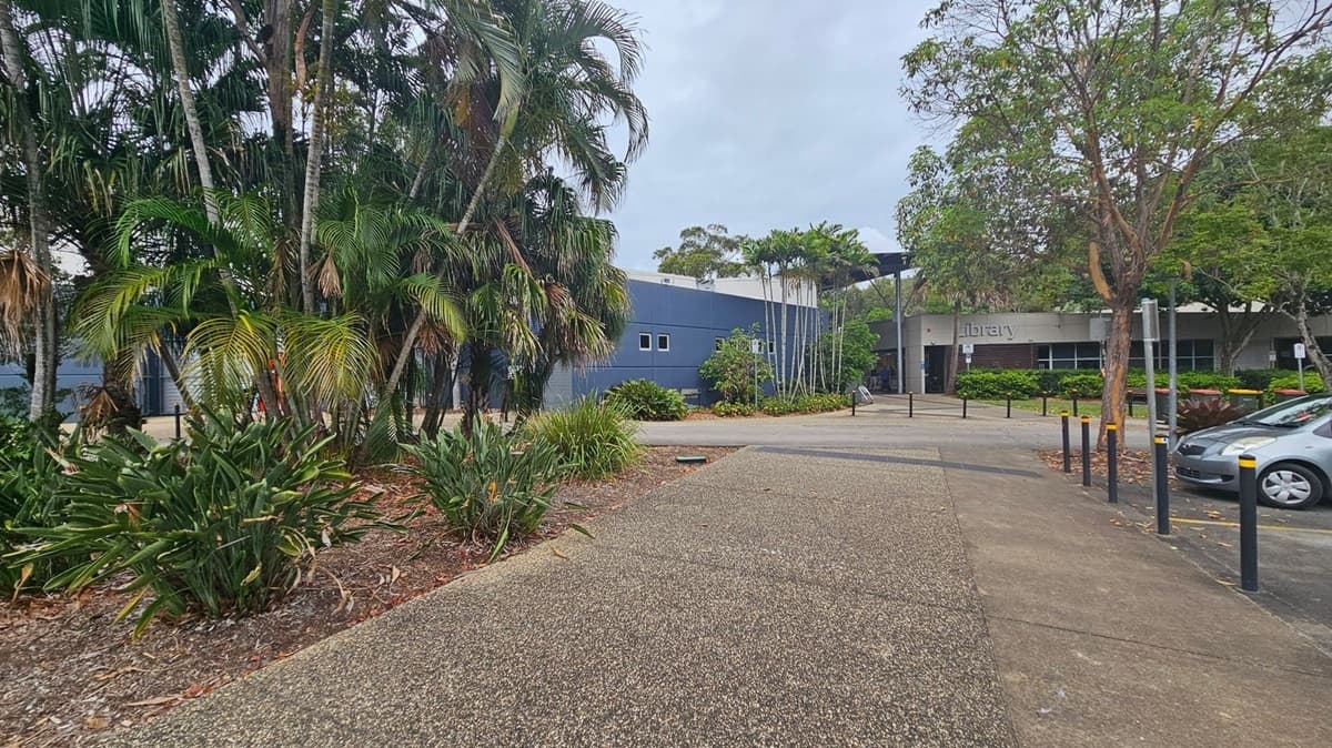 A pathway leads towards a building with a sign that reads "library" on the right. The building is surrounded by lush greenery, including tall palm trees and various shrubs. On the left, there is a dense cluster of palm trees and plants. A car is parked on the right side near the building. The ground is covered with a textured pathway and some concrete. The sky is overcast, and there are bollards lining the path.