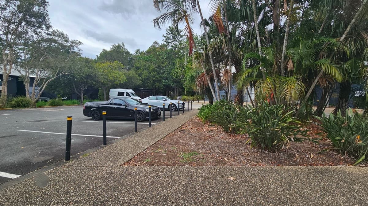 A car park with several vehicles, including a black car and a white van, is on the left. The ground is paved with a textured surface. On the right, there are tall palm trees and dense green foliage. Black bollards with yellow tops line the edge of the pavement. In the background, more trees and greenery are visible, creating a lush setting.