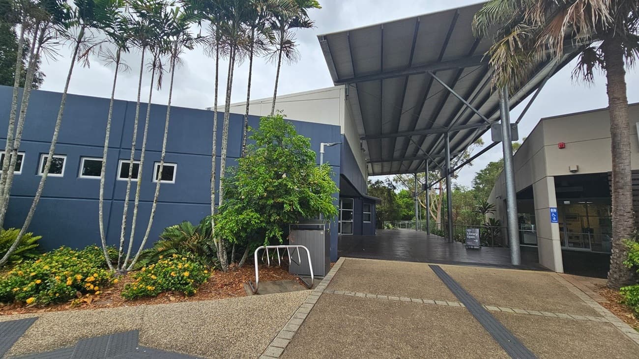 A modern building with a large, overhanging metal roof supported by steel beams. On the left, there is a blue wall with small square windows and tall, slender palm trees in front. Yellow flowers and green shrubs are planted along the base. In the centre, a bike rack is visible. On the right, an entrance with a sign indicating the library is open. The ground is paved with textured tiles, and there are accessible signs near the entrance.